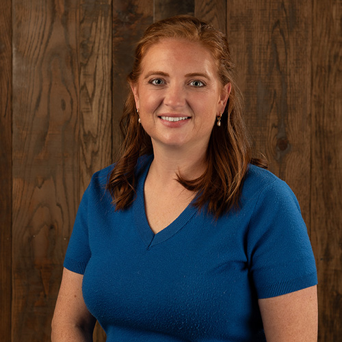 Portrait of a smiling woman in a blue short-sleeve shirt, standing against a wooden panel background.