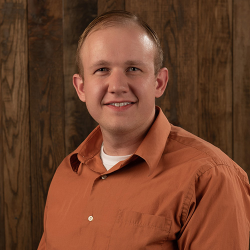 Portrait of a smiling man in an orange button-down shirt against a wooden backdrop.