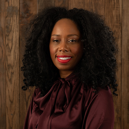 Portrait of a smiling woman with curly black hair, wearing a burgundy blouse with a bow, against a wooden backdrop.