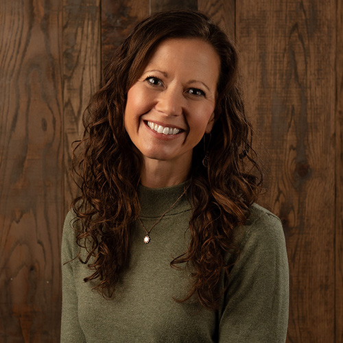 Portrait of a smiling woman with long wavy brown hair wearing a green sweater, against a wooden panel background.