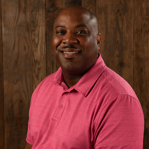 Portrait of a smiling man in a pink polo shirt against a wood-paneled backdrop.