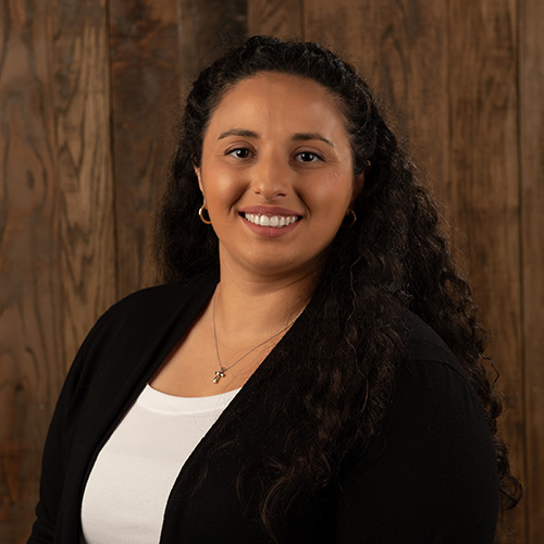 Smiling woman with long curly hair in a black cardigan and white top, standing against a wooden backdrop.
