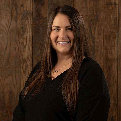 Smiling woman with long brown hair, wearing a black top, posed against a wood-paneled background.