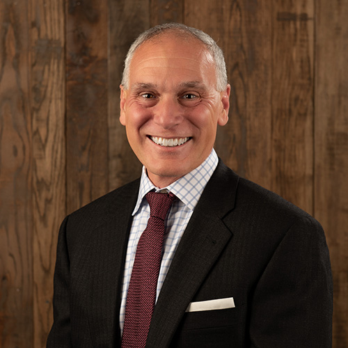 Professional headshot of a smiling man in a dark suit, white shirt, and red tie, standing against a wooden backdrop.