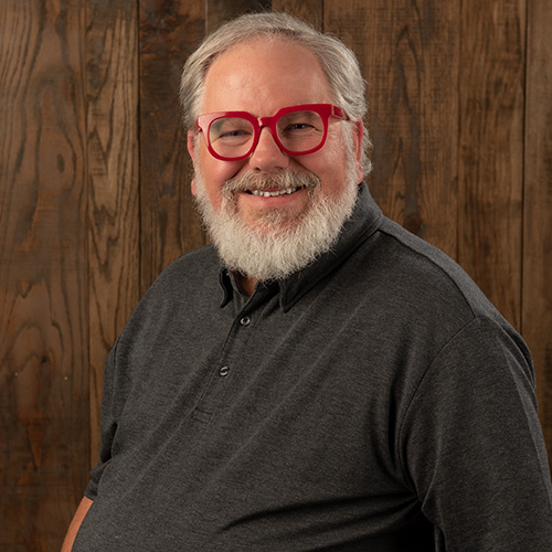 Smiling man with gray beard and red glasses in a dark polo, standing against a wooden backdrop.
