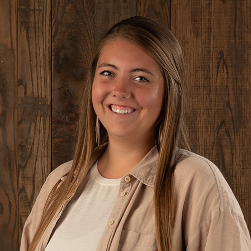 Smiling woman with long light-brown hair, wearing a beige button-up shirt, standing in front of a wooden paneled background.