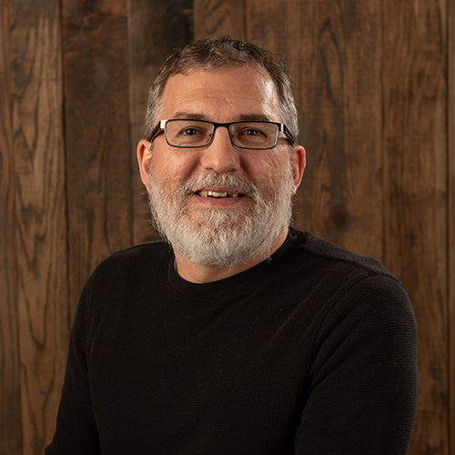 Portrait of a smiling man with gray beard and glasses wearing a black sweater, standing against a wooden panel background in a studio setting.