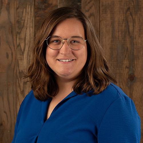 Smiling woman with round glasses in a blue blouse, standing against a wooden background.