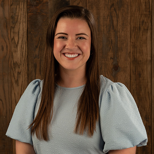 Smiling woman with long brown hair wearing a light blue blouse, standing in front of a wooden wall.