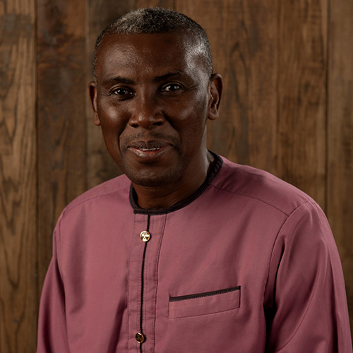 Portrait of a Black man in a mauve collared shirt with a small chest pocket, standing against a wooden backdrop.