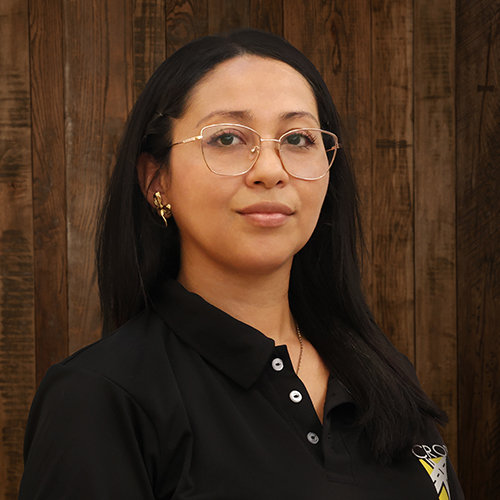 Portrait of a woman with long dark hair and gold-rimmed glasses, wearing a black polo, against a wooden paneled background.