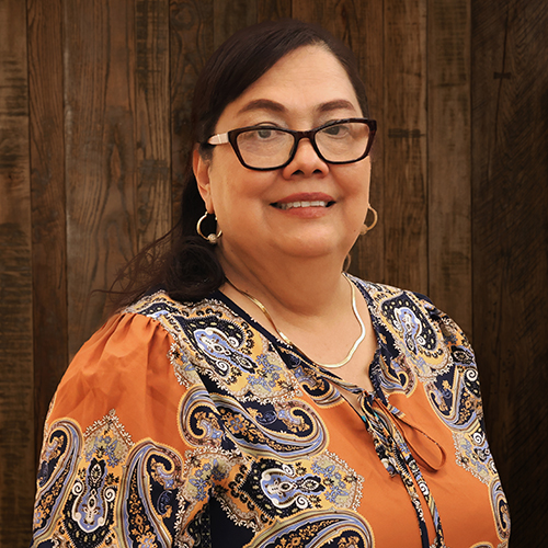 Smiling woman with dark hair and glasses, wearing an orange paisley blouse, standing against a wooden wall.