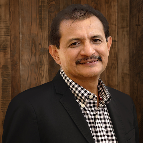 Smiling man in a black blazer and checkered shirt posing against a wooden wall background.
