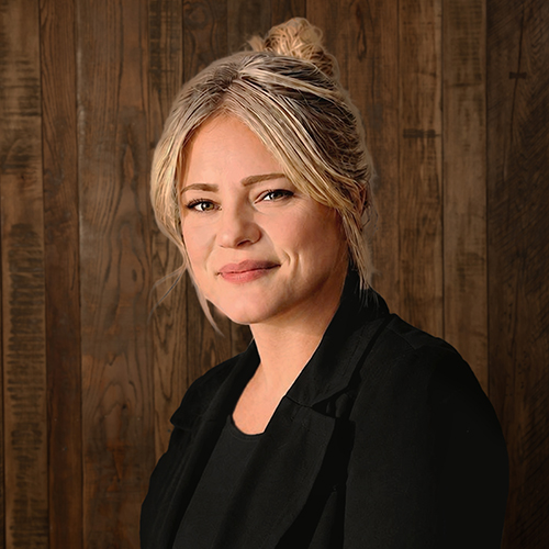Portrait of a smiling woman with blonde hair in a bun, wearing a black blazer, standing against a wood-paneled wall.