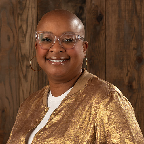 Smiling bald Black woman wearing clear glasses and a metallic gold jacket, posed against a wooden backdrop.