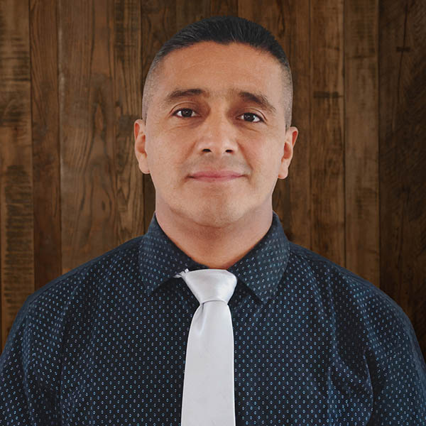 Portrait of a man in a dark dotted shirt and white tie, standing against a wood-paneled backdrop.