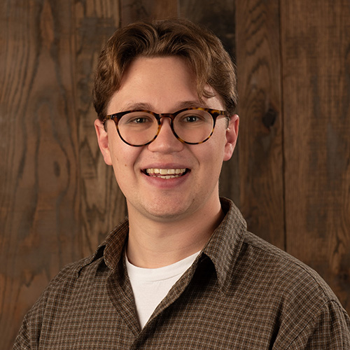 Smiling person with short brown hair and tortoiseshell glasses, wearing a brown plaid shirt, posed against a wooden backdrop.